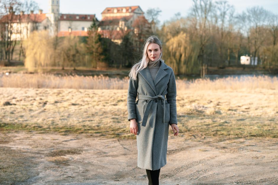 A woman in a grey coat poses elegantly outdoors near Нясвiж Palace in Belarus.