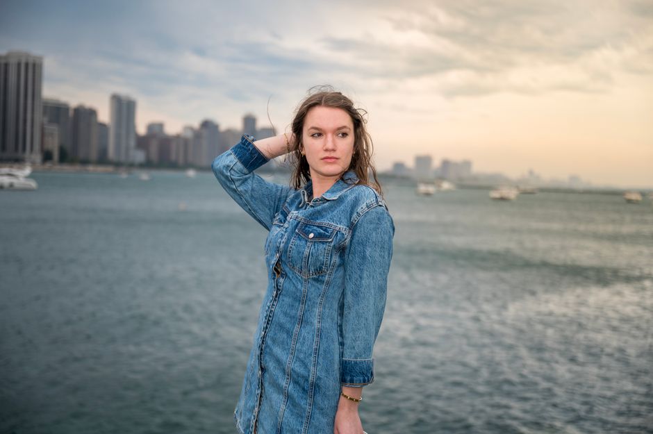 A woman in denim poses by a city waterfront with skyscrapers in the background under a cloudy sky.