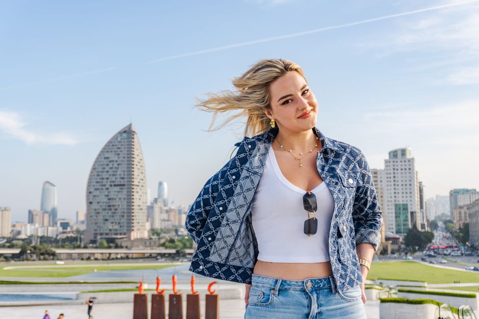 Casually dressed young woman smiles in front of a modern cityscape with iconic buildings in the background.