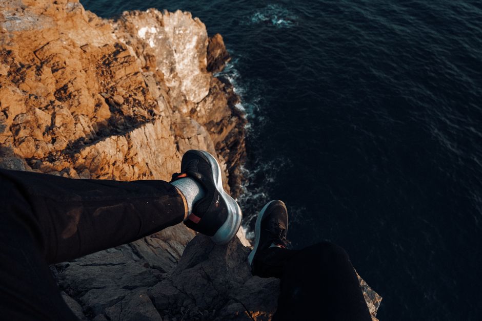 A dramatic POV of feet dangling over a rocky ocean cliff with waves.