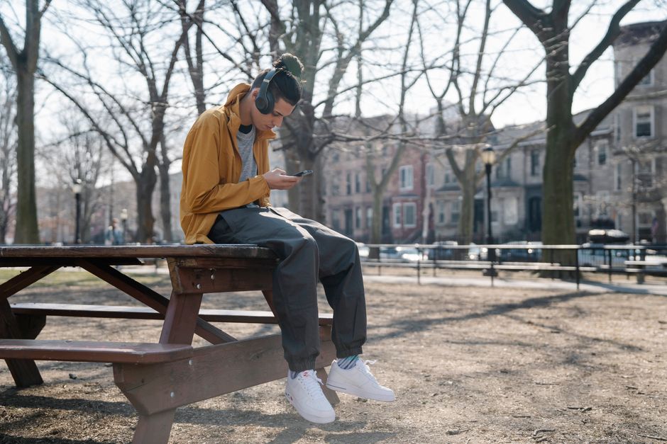 Cool ethnic male in headphones text messaging on cellphone while listening to music and sitting on table in town
