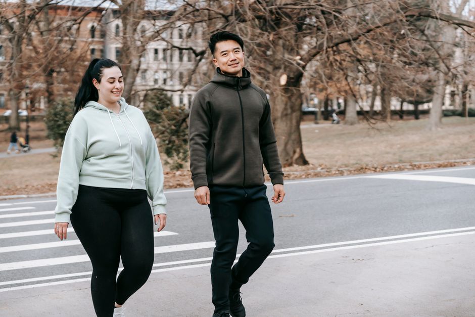 Positive young multiethnic male and female friends in sportswear smiling while crossing asphalt road near city park after outdoor training