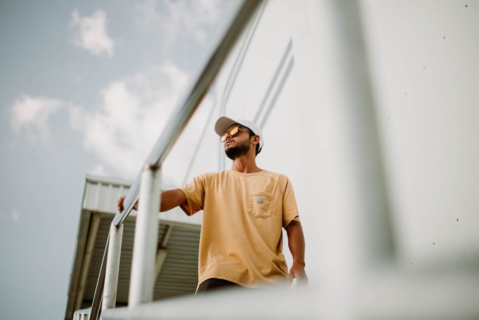 Confident man with sunglasses and cap standing by railings in modern urban setting, captured in sunlight.