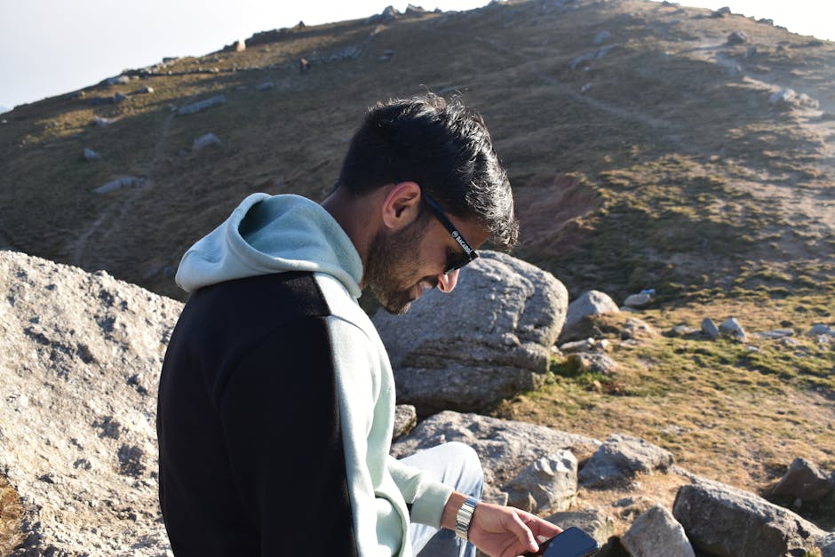 A young man relaxes on a rocky hillside, enjoying the outdoors with a smartphone.