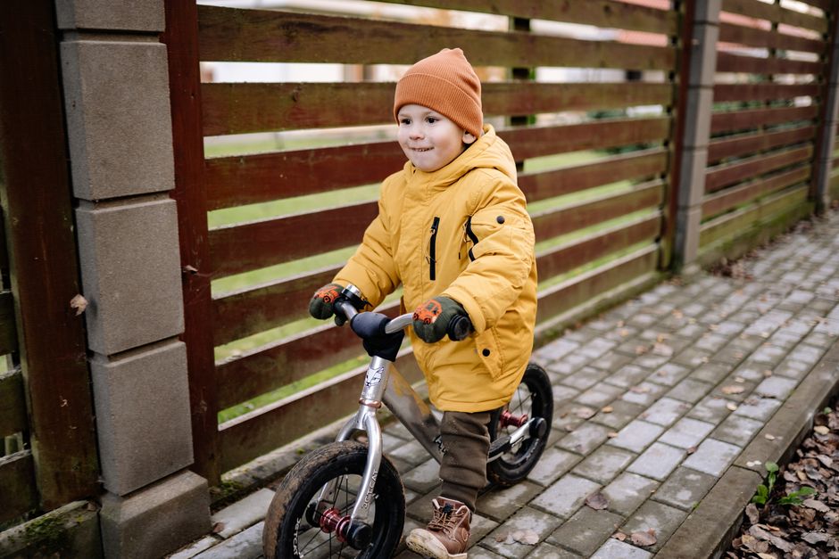 A cheerful child wearing a yellow jacket rides a balance bicycle along a paved walkway.
