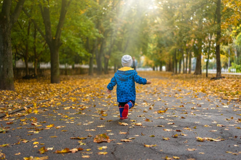 A young child in a blue jacket runs joyfully through autumn leaves in a sunny park.