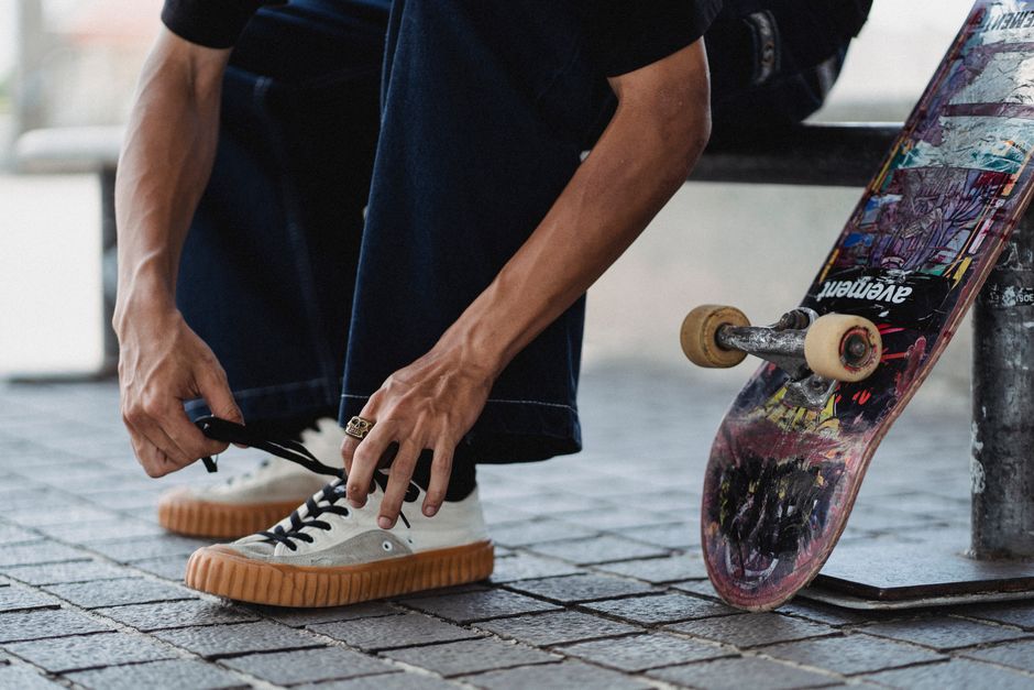 Crop unrecognizable male skater sitting on bench while tying shoelaces on white sneakers before training on skateboard