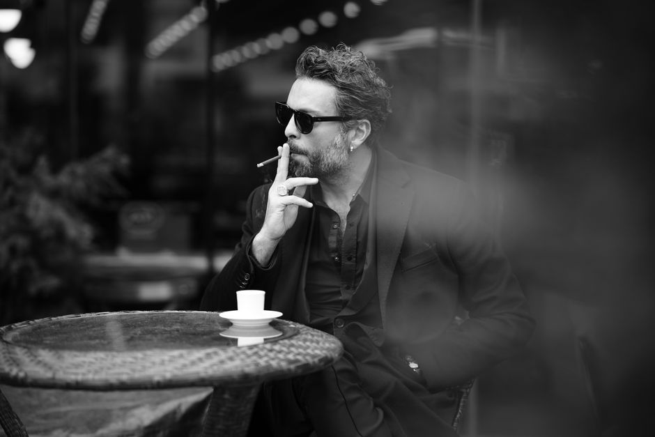 Monochrome image of a man in a black suit smoking at a cafe, exuding sophistication.