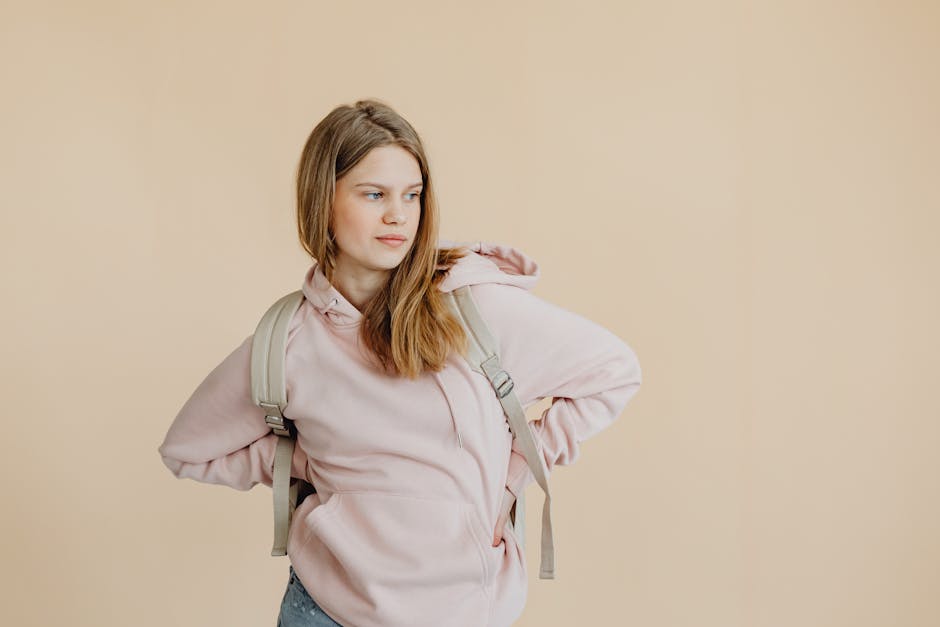 Portrait of a young woman in a pink hoodie and backpack against a beige background.