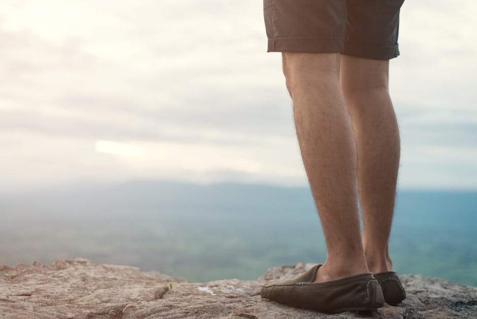 Close-up of a man's legs in casual shoes standing on a rocky edge overlooking a scenic landscape.