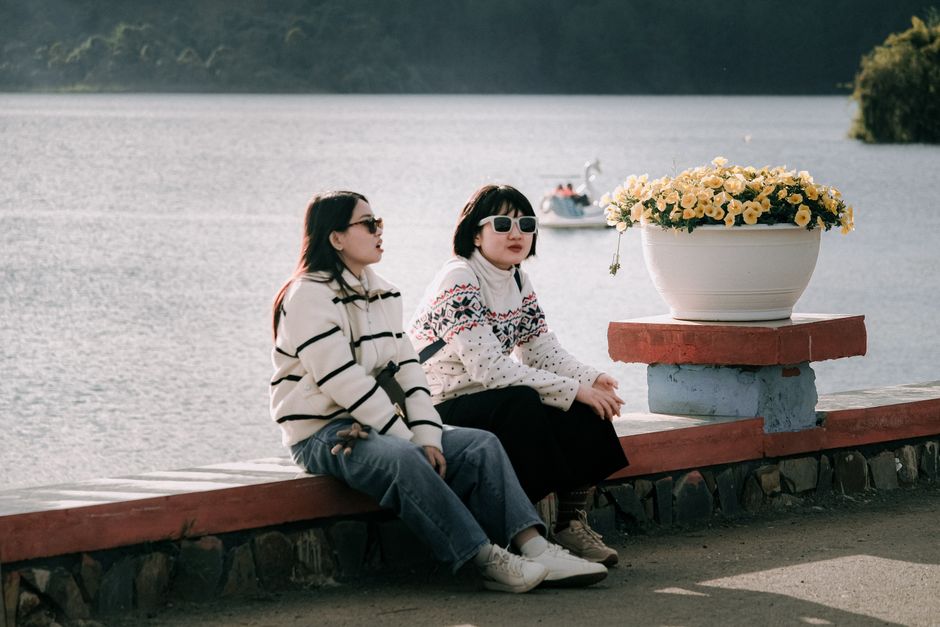 Two women in knit sweaters and sunglasses sitting by a serene lake with flowers.