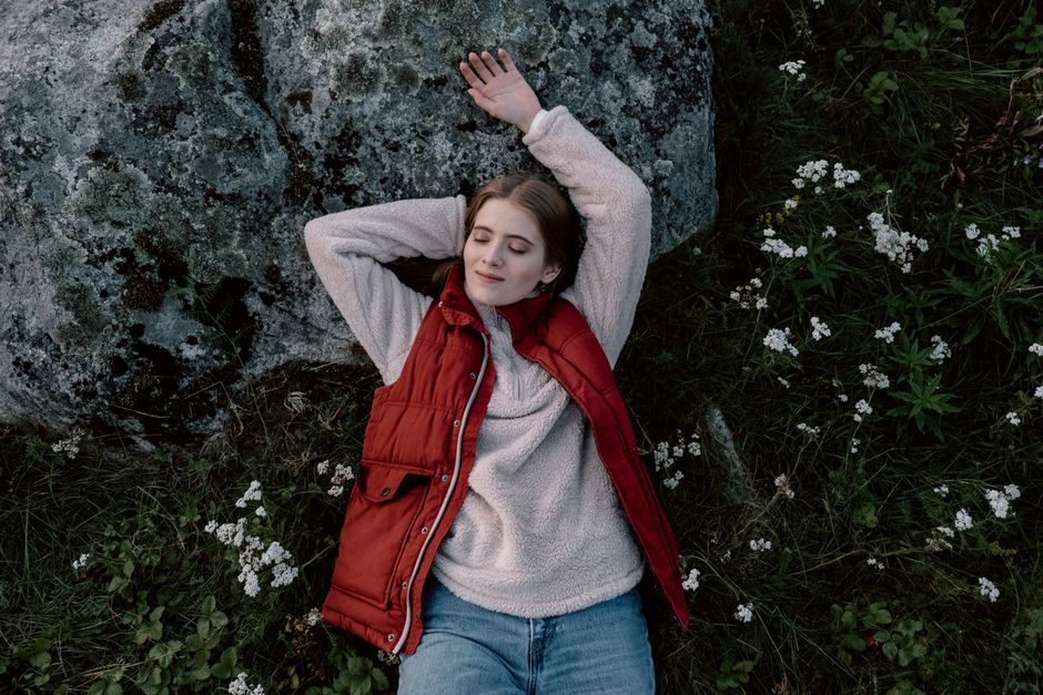 A serene scene of a young woman lying outdoors on grass, eyes closed in relaxation, surrounded by flowers.