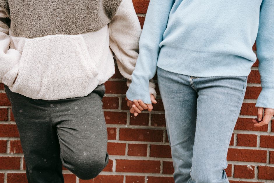 Crop unrecognizable couple in warm sweaters standing near brick wall of building and holding hands in snowy day