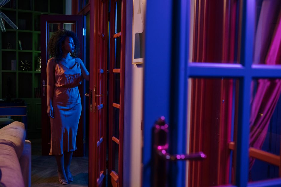 A woman in a satin dress stands by an open doorway at night lit in colorful tones.