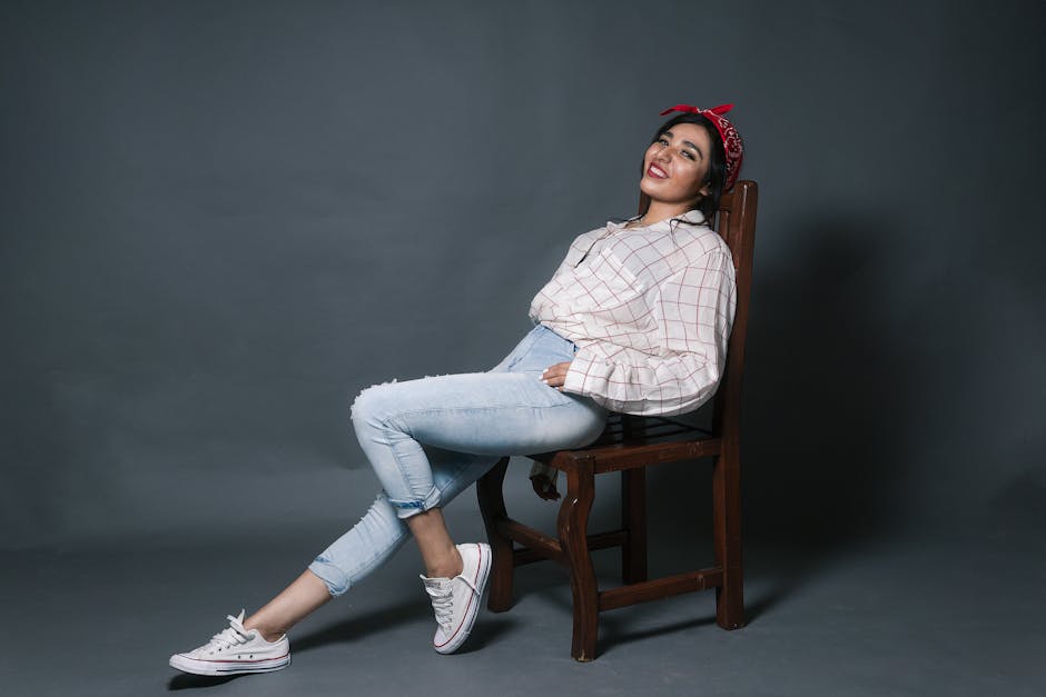 Fashionable woman in casual attire, posing confidently on a chair with a gray backdrop.