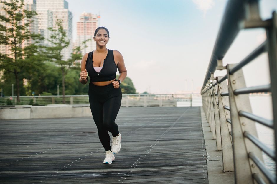 A woman in activewear jogging on a boardwalk by the waterfront with city skyscrapers in the background.