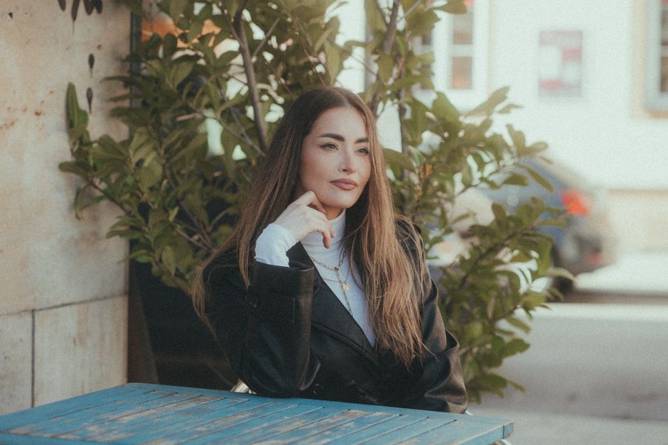 Woman in a leather jacket sitting at a table, posing outdoors with plants in the background.