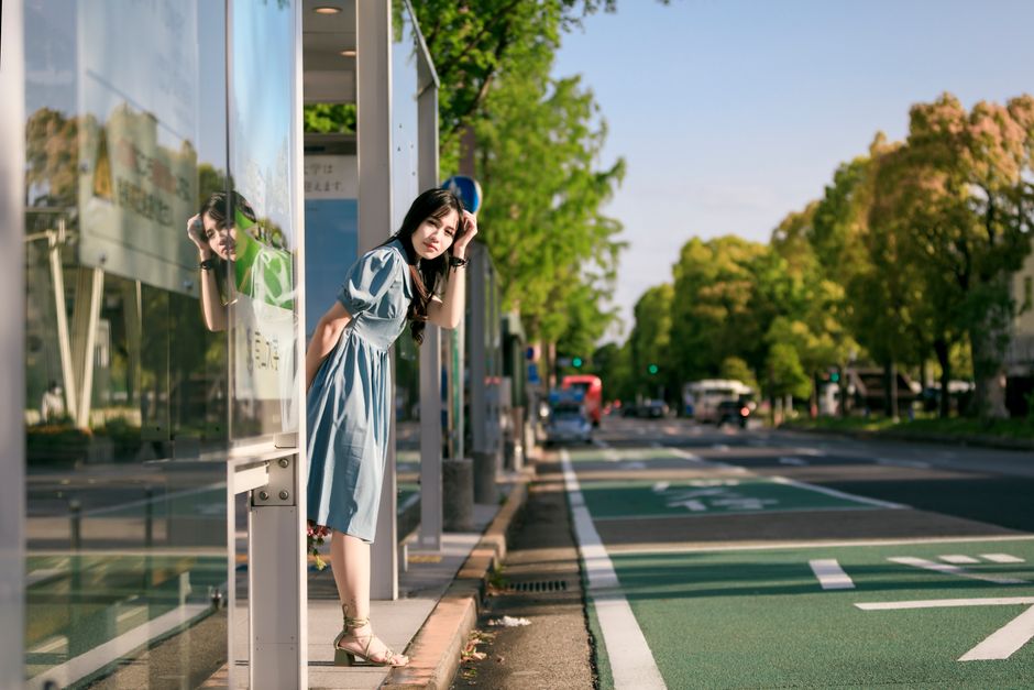 An Asian woman stands at a bus stop on a sunny day, looking down the street.