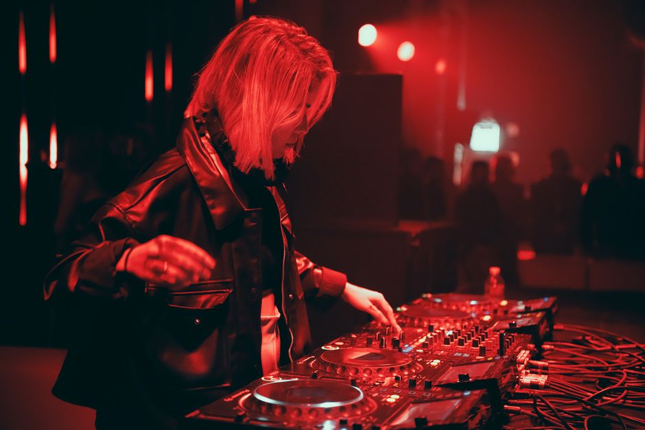 A talented DJ in a leather jacket playing electronic music at a lively nightclub.