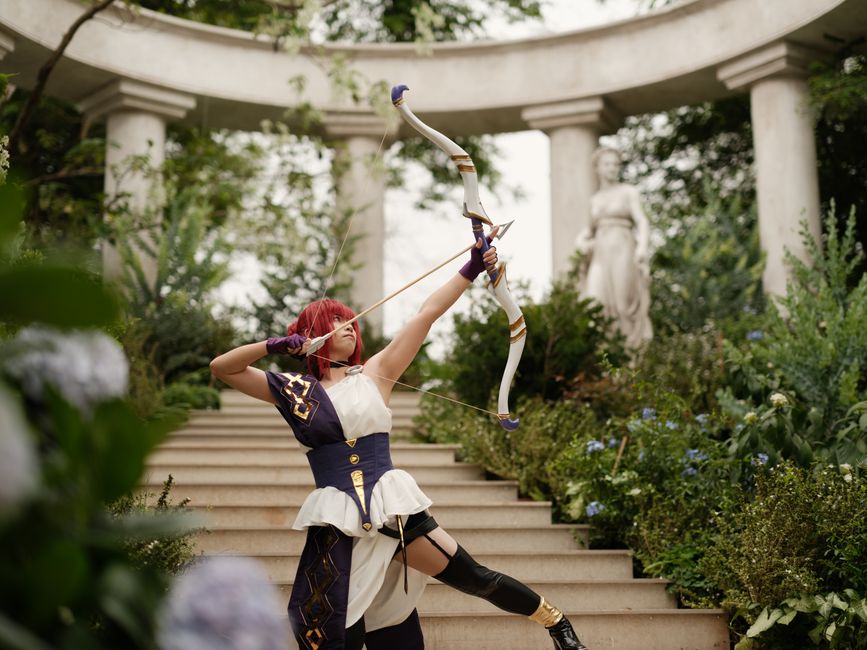 Cosplayer in elaborate costume practices archery in a lush garden setting, capturing a sense of fantasy and elegance.