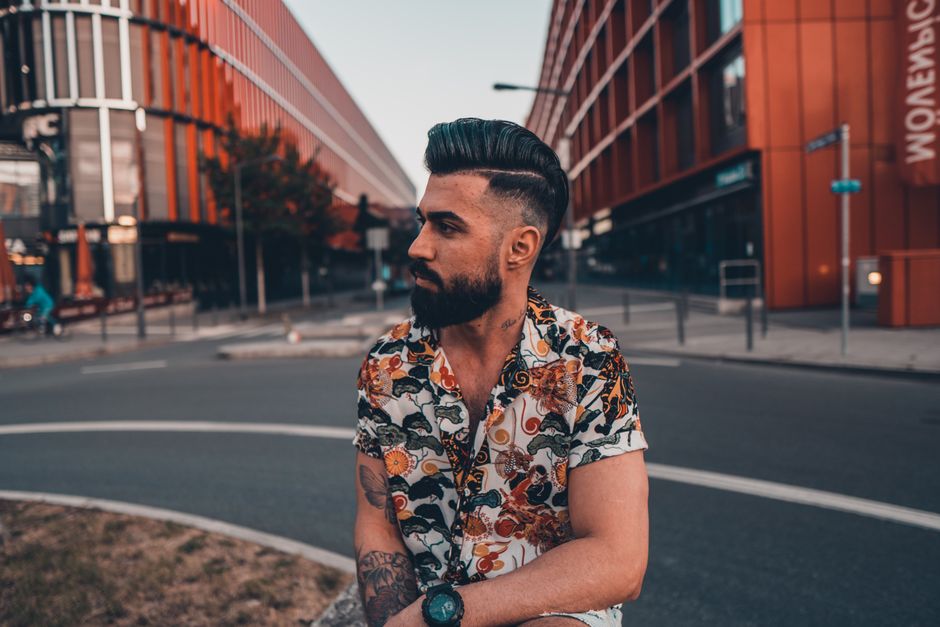 Confident bearded pensive male in trendy colorful casual shirt with stylish haircut resting on city street with modern building in urban area