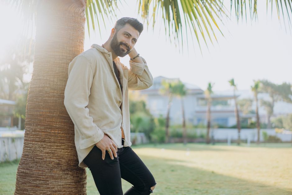 Stylish man smiling while leaning on a palm tree in a sunny outdoor garden setting.