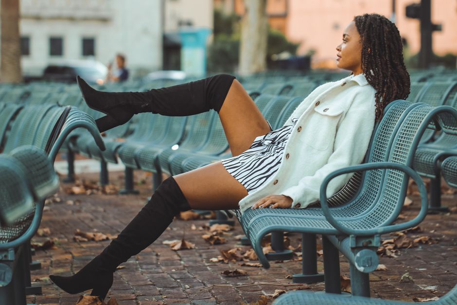 Fashionable woman in boots and white jacket lounging on a metal bench surrounded by fallen leaves.