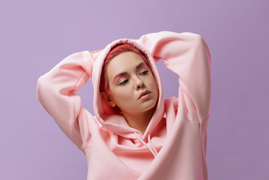 Studio portrait of a woman with pink hair and hoodie against a lilac background.